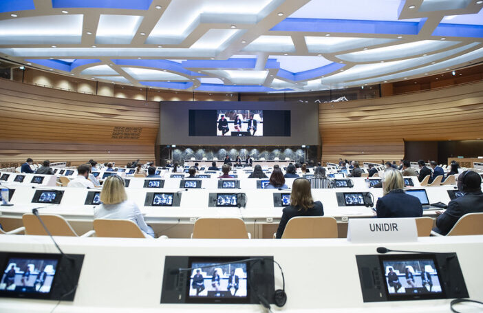 Photograph of a large conference room where all all delegates have a small video screen and headphones and all are looking at large screen with three people seated on stools.