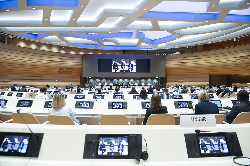 Photograph of a large conference room where all all delegates have a small video screen and headphones and all are looking at large screen with three people seated on stools.