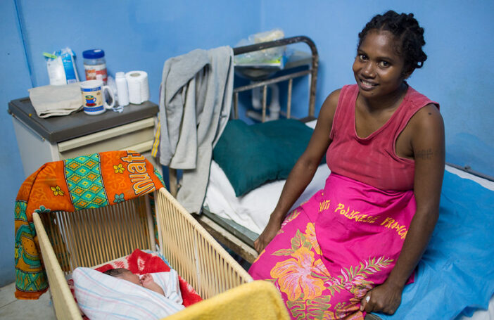 Photograph of a smiling mother sitting on a bed beside a cradles with a new-born baby