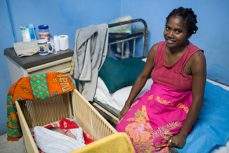 Photograph of a smiling mother sitting on a bed beside a cradles with a new-born baby