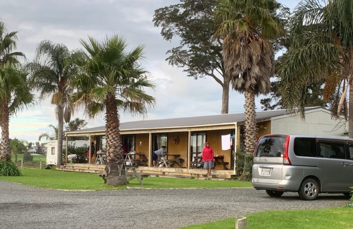 Photograph of a single story building with a verandah on which there are three people and some outdoor tables and benches. There are tall palm trees growing around the house and a small van parked in the driveway.