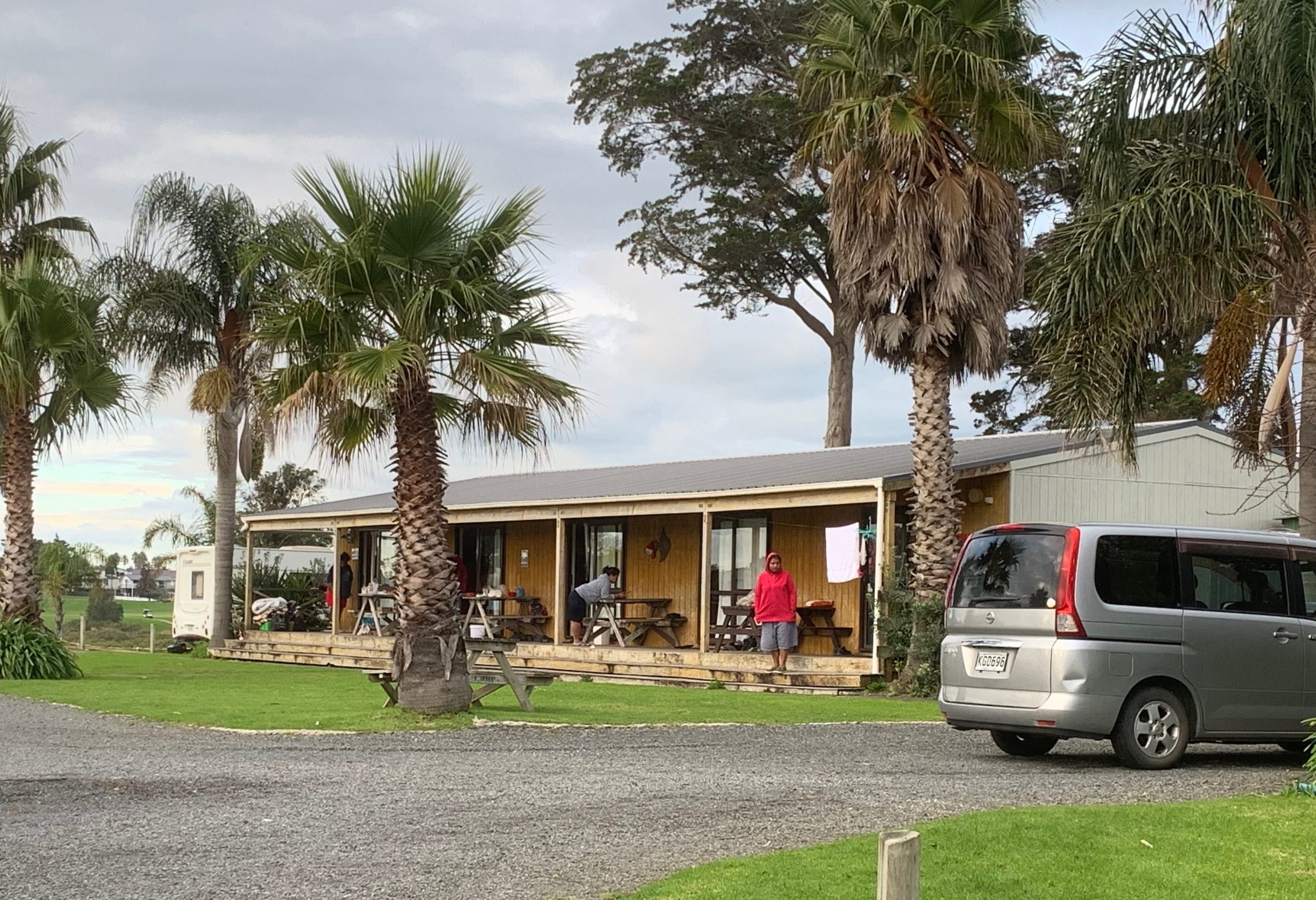 Photograph of a single story building with a verandah on which there are three people and some outdoor tables and benches. There are tall palm trees growing around the house and a small van parked in the driveway.