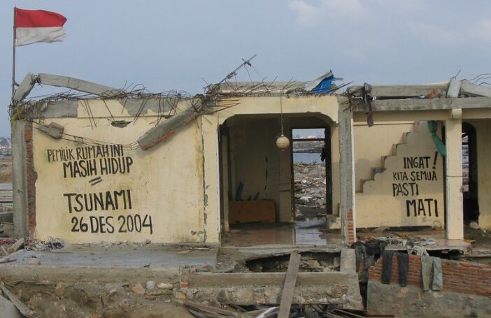 A photograph of a cream coloured building that was partially destroyed by a tsunami in 2004. Large letters hand-written on the walls say "Pemuk rumahini masih hidup. Tsunami 26 Des 2004. Ingat! Kita semua pasti mati." A red and white flag flies on the roof.