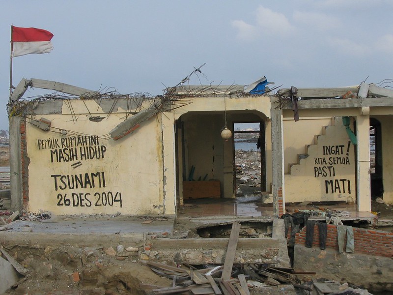 A photograph of a cream coloured building that was partially destroyed by a tsunami in 2004. Large letters hand-written on the walls say "Pemuk rumahini masih hidup. Tsunami 26 Des 2004. Ingat! Kita semua pasti mati." A red and white flag flies on the roof.