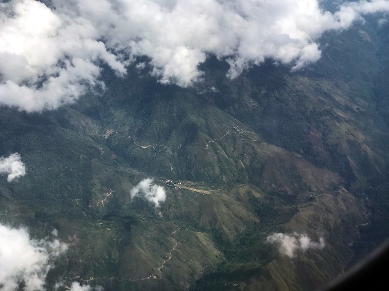 An aerial view of the highlands of Papua New Guinea. There are clouds in the foreground and far below buildings and narrow roads.