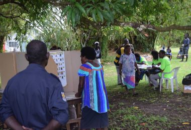 A woman in a colourful top and dark blue skirt is looking at a poster of election candidates in an outdoor polling station in Bougainville, PNG.