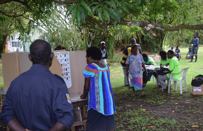 A woman in a colourful top and dark blue skirt is looking at a poster of election candidates in an outdoor polling station in Bougainville, PNG.