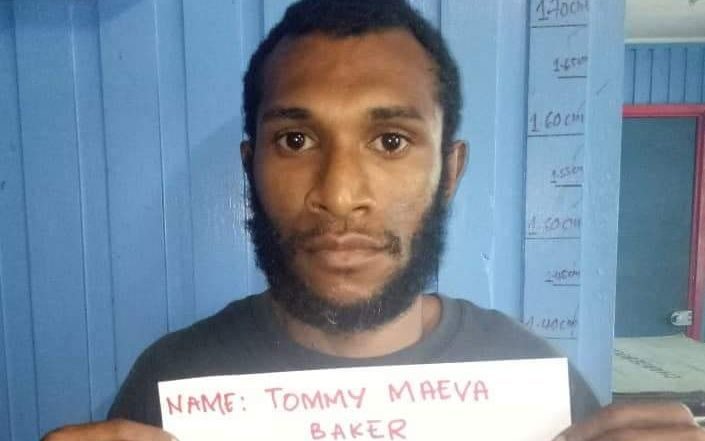Photograph of head and shoulders of a man with dark skin, black hair and black beard. He is in front of a blue wall with height markings written on the doorway edge. He is holding a white sign with hand writing in red that says 'Name: Tommy Maeva Baker'.