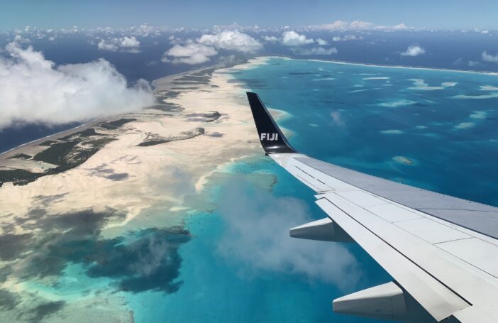 Image shows the view from an airplane over the wing. There are clouds in the mid distance and below there is the ocean in varying shades of blue, golden sands, and green vegetation.