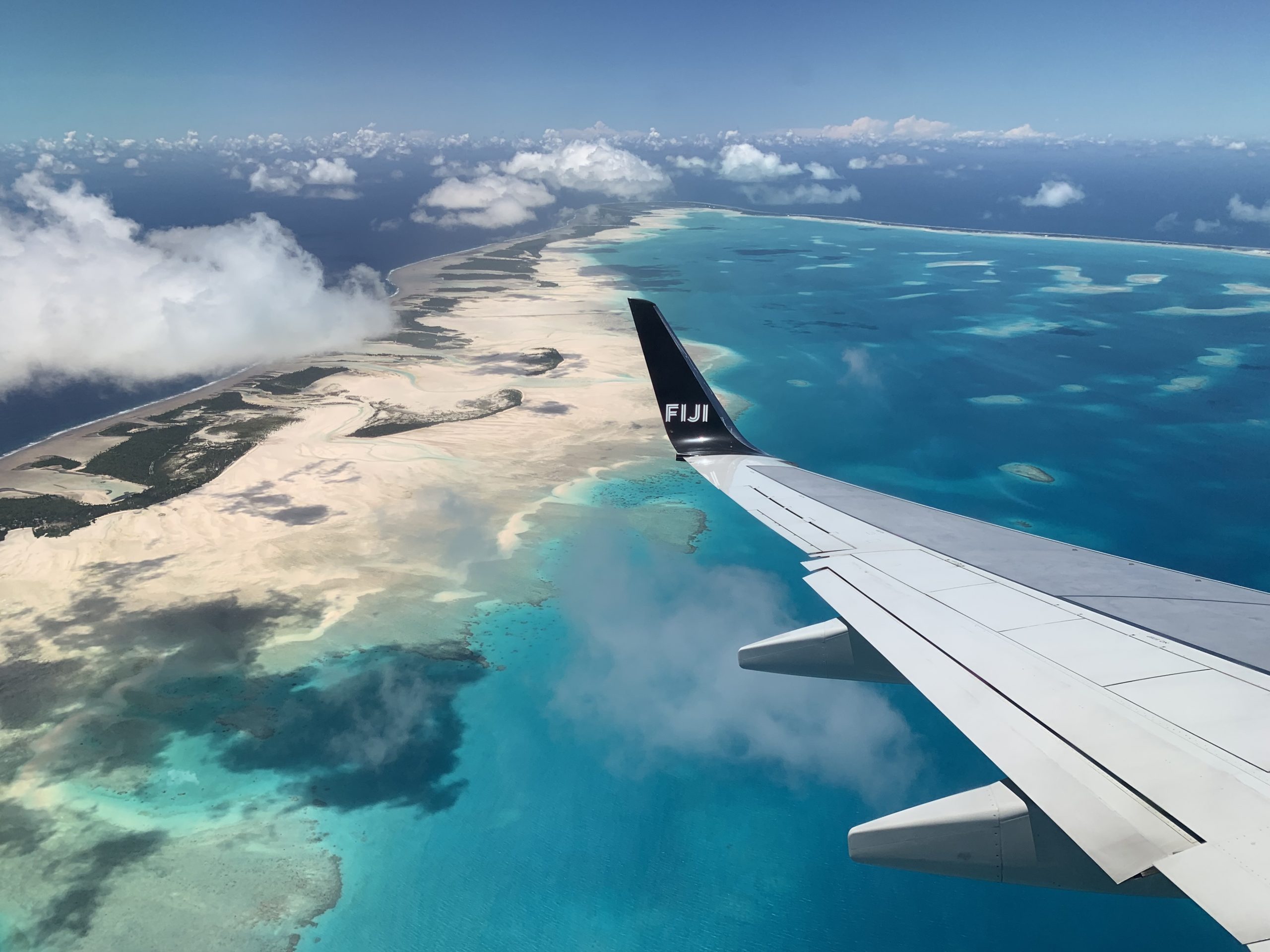 Image shows the view from an airplane over the wing. There are clouds in the mid distance and below there is the ocean in varying shades of blue, golden sands, and green vegetation.