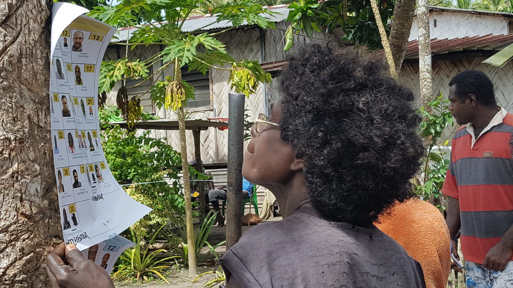 A women in Bougainville, Papua New Guinea, checks the photos and descriptions of candidates in the 2017 elections. The notice is attached to a tree. The woman has black curly hair and wears glasses. In the background is a thatched hut, a garden, and a man in a red and grey striped t-shirt.
