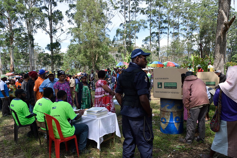 Photograph of people voting in Papua New Guinea. Voters in colourful clothing queue in front of a table where three election officals in high-vis shirts sit. A policeman stands on the grass in the foreground and there are tall trees in the background.
