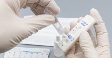 Photograph of rubber-gloved hands, one hand is squeezing a sample from a soft plastic bottle onto a COVID-19 antigen test stick held in the other hand.