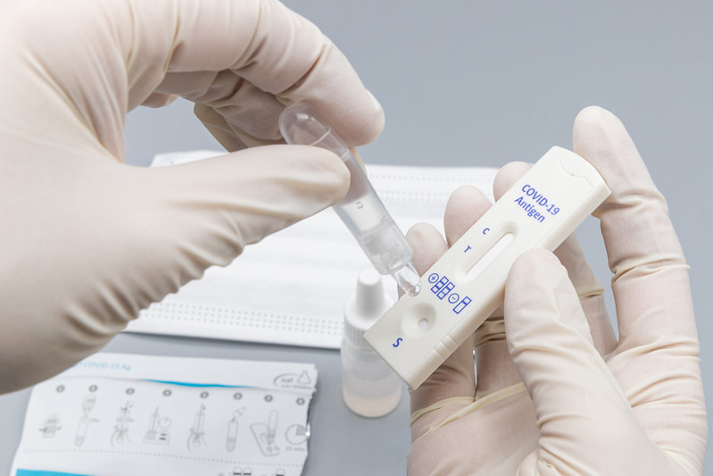 Photograph of rubber-gloved hands, one hand is squeezing a sample from a soft plastic bottle onto a COVID-19 antigen test stick held in the other hand.