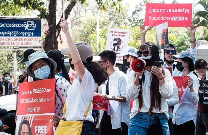 Photograph of people at a February 2021 protest agains the military coup in Myanmar. People are wearing masks and carrying signs. One woman holds a megaphone to her masked mouth.