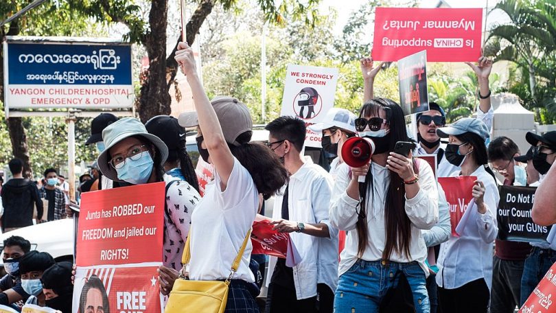 Photograph of people at a February 2021 protest agains the military coup in Myanmar. People are wearing masks and carrying signs. One woman holds a megaphone to her masked mouth.