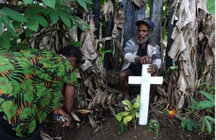 Photograph of a man and a woman clearing the area around a grave, marked by a white cross, among dense trees and bushes.