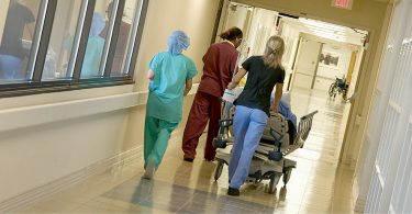 Photograph of a hospital corridor: ahead three hospital staff accompany a patient being moved on a bed.