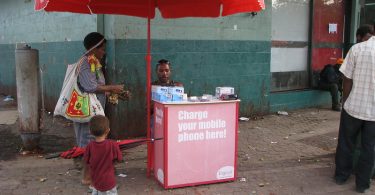 A woman and a child talk to a man sitting under a Digicel-branded umbrella behind a small booth with signage that says "Charge your mobile phone here".