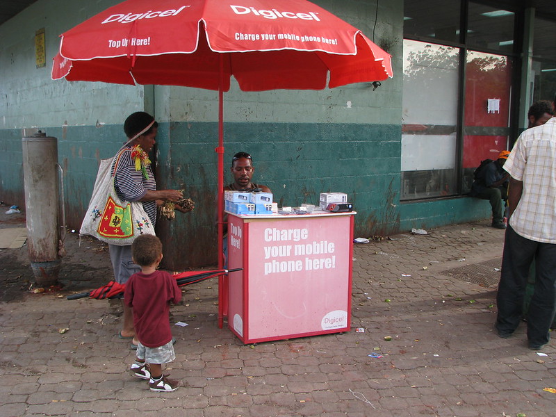A woman and a child talk to a man sitting under a Digicel-branded umbrella behind a small booth with signage that says "Charge your mobile phone here".