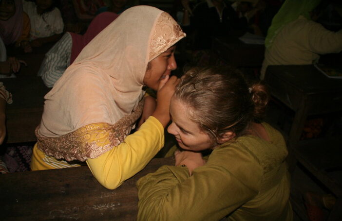 A young woman in a yellow dress and pale peach head scarf is whispering into the ear of a young woman with blonde hair wearing a khaki green shirt.