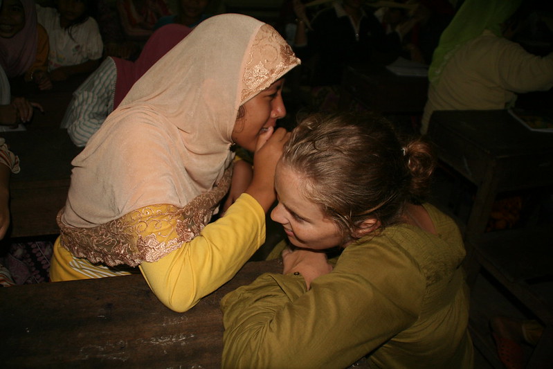 A young woman in a yellow dress and pale peach head scarf is whispering into the ear of a young woman with blonde hair wearing a khaki green shirt.