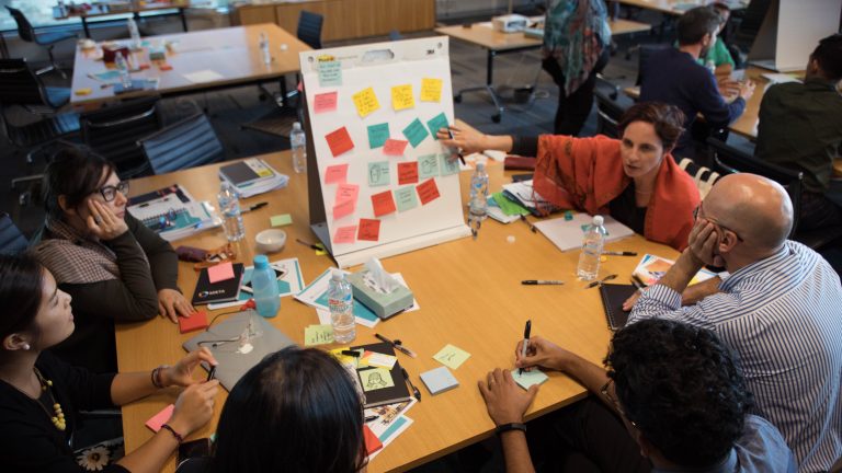 People sit around a table with a white board and coloured post-it notes at an IRF "boot camp" session.